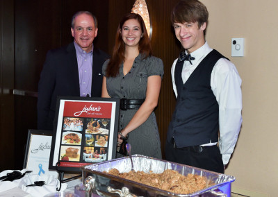 Three people standing at a catering event, one holding a framed advertisement, with a table featuring a dish in the foreground.