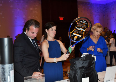 Three adults examine silent auction items at a charity event; one holds a tennis racket.