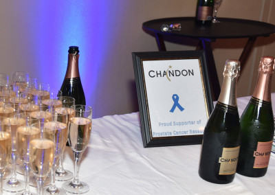 Event table with chandon sparkling wine bottles, filled champagne glasses, and a sign supporting prostate cancer research.