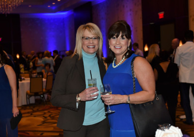 Two women smiling at a social event, holding drinks, with one in a blue dress and the other in a turquoise turtleneck, in a room with blue lighting.