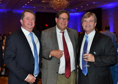 Three men in business attire, with one holding a wine glass, smiling and posing together at a formal event.