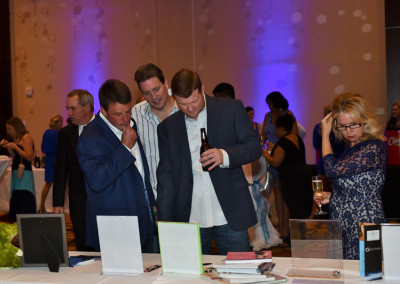 Three men examine papers at a table during an event, with other attendees in background, ambient lighting, and a festive atmosphere.