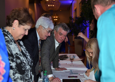 Guests at a formal event lean over a registration table, examining documents and talking to a young woman seated behind the table.