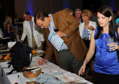 A man in a brown blazer signs a document at a table during an event, as a woman holding a drink looks on.
