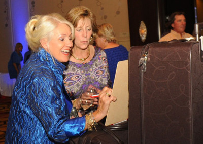 Two women, one in a blue blouse and the other in a purple patterned top, are looking at a laptop screen together at an event.