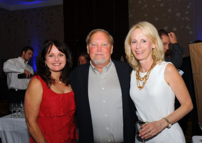 Three adults posing at a social event, with a woman in a red dress on the left, a man in a blue shirt in the center, and a woman in a white dress on the right.