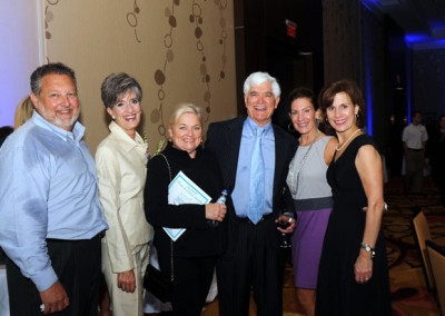 Six smiling adults standing together at a formal event, with one man holding a book and a woman holding a glass of wine.