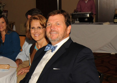 A smiling man with a beard and bow tie seated next to a woman at a formal event, with other guests and a table in the background.