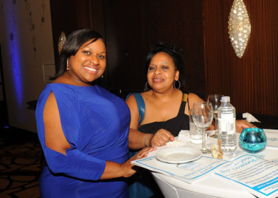 Two women smiling at a table during a formal event, with a dinner plate in front of them and decorative items on the table.