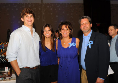 Family of four smiling at a formal event, all wearing blue ribbons, with a bokeh background.