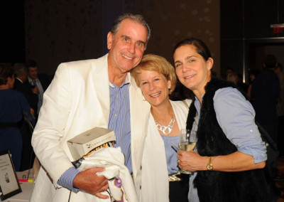 Two women and a man smiling at a social event, one holding a wine glass and another carrying a white jacket.
