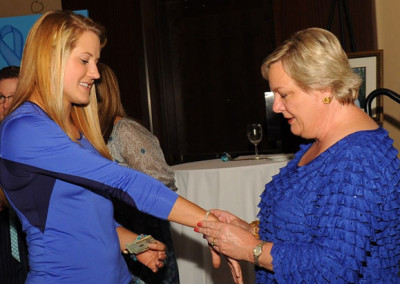 Two women shaking hands at an event, one in a blue dress and the other in a blue blouse, both smiling.