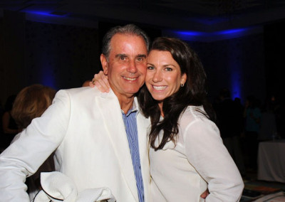 An older man in a white suit and a younger woman in a cream jacket smiling together at a social event.