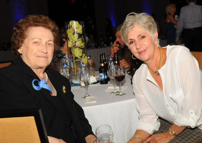 Two elderly women sitting at a table during an event, smiling at the camera with drinks and floral centerpiece visible.