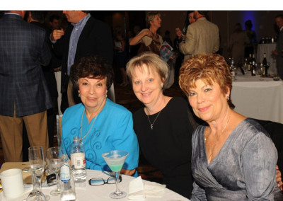 Three women smiling at a table during a social event, with other guests mingling in the background.