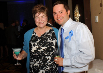 A woman and a man smiling at a formal event, holding drinks; the woman in a floral black and white dress and the man in a blue-striped shirt with a ribbon pin.