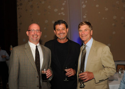 Three smiling men in suits holding drinks at a formal event.