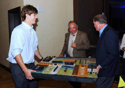 Young man presenting a colorful architectural model to two older men in a formal setting.