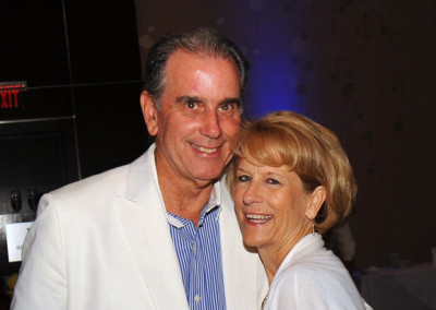 An older man and woman smiling at a social event, the man in a white suit and the woman in a grey cardigan.