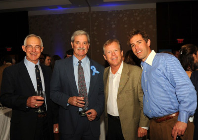 Four men smiling at a social event, holding wine glasses with a blue ribbon pinned on one man’s jacket.