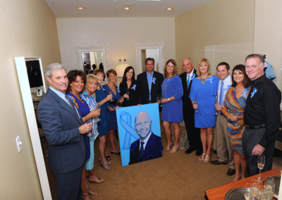 Group of adults in a room holding wine glasses and a large portrait of a man, smiling and wearing blue ribbons.