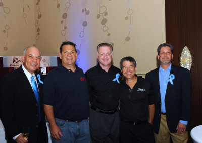 Five men wearing blue ribbons posing together at a charity event.
