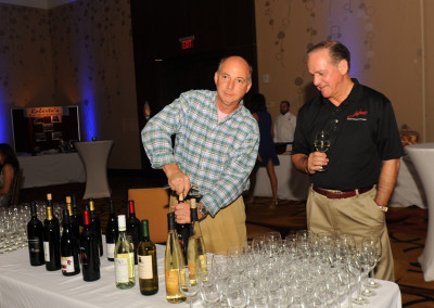 Two men at a wine tasting event, one pouring wine and the other holding a glass, with bottles and glasses arranged on a table.
