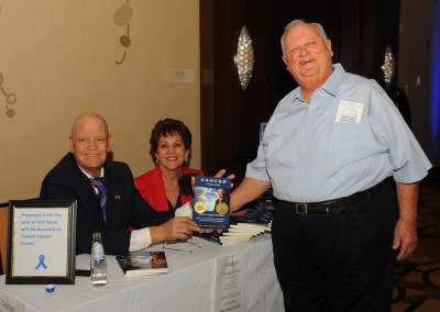 Three people at a book promotion event, with one man standing and holding a book titled "cancer" while the seated couple smiles at the camera.