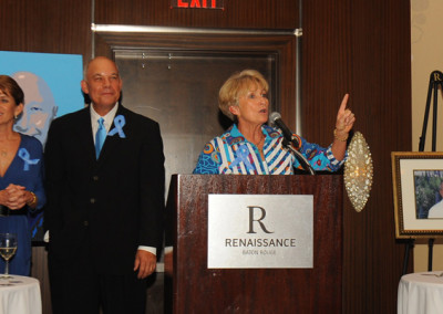 Three people at a podium during an event, with one woman speaking and two listening, surrounded by blue ribbons and a portrait on an easel.