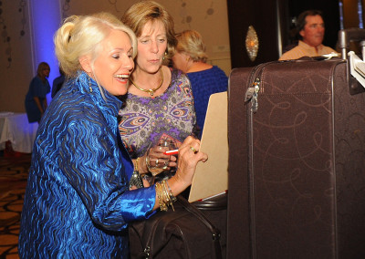 Two women examining items at a silent auction table during an event, with other attendees in the background.