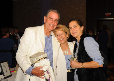 Three people smiling at a social event; two women and one man, all dressed in semi-formal attire, standing closely together in a room with other attendees in the background.