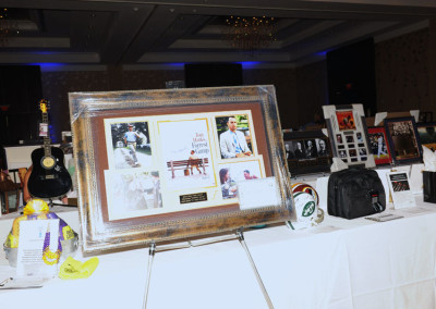 A display table at an event featuring a large framed collage of photos and memorabilia, including a guitar and backpack.