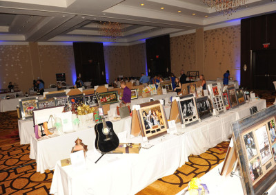 Ballroom set up for an auction with tables displaying items such as framed pictures, a guitar, and sculptures under chandeliers.