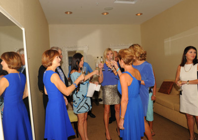 Group of women in a room, some in blue dresses, interacting and holding drinks, with one woman in a sequined skirt.