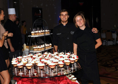 Two caterers, a man and a woman, standing beside a dessert table with cupcakes and small treats at an event.