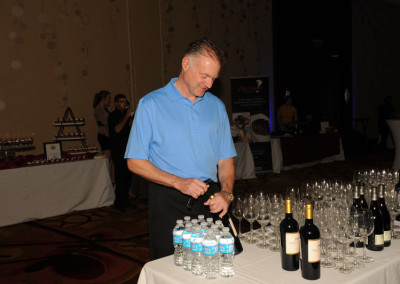 A man in a blue shirt opening a bottle at a beverage table with wine bottles and water bottles during an event.