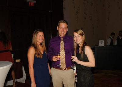Three people posing for a photo at a formal event, two women and a man in the center holding drinks, with a dimly lit background.