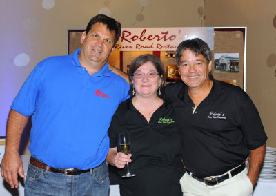 Three adults smiling at a promotional event for roberto's river road restaurant, standing in front of a banner.