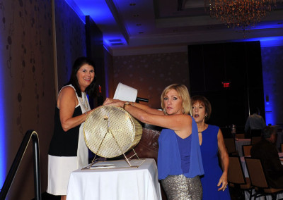 Three women at a formal event, one drawing a raffle ticket from a large lottery drum.