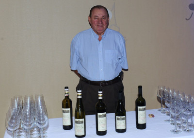 Man standing behind a table with five bottles of wine and rows of empty wine glasses.