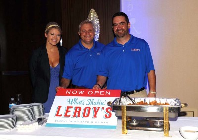 Three smiling people in blue shirts stand behind a promotional sign for leroy's restaurant at an indoor event.