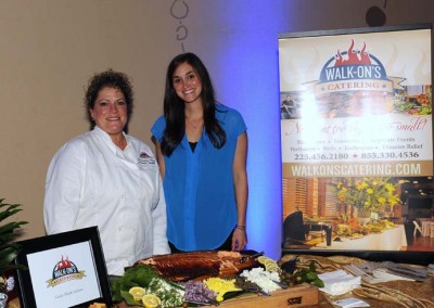 Two women, one in a chef's coat and another in a blue polo shirt, stand beside a catering display table under a "walk-on's catering" sign.