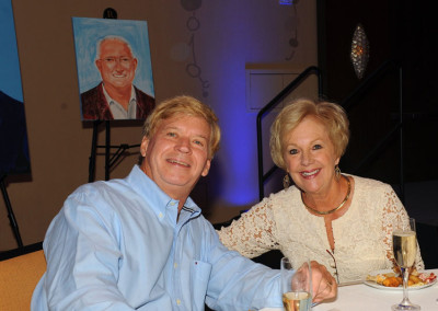 An older couple smiling at the camera while sitting at a table during an event, with a portrait painting displayed in the background.