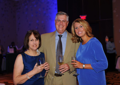 Three adults at a formal event, holding champagne glasses and smiling at the camera, with a dimly lit, blue-lit background.