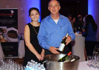 Two people, a man and a woman, standing at a wine tasting event with a bottle of wine and a bucket.