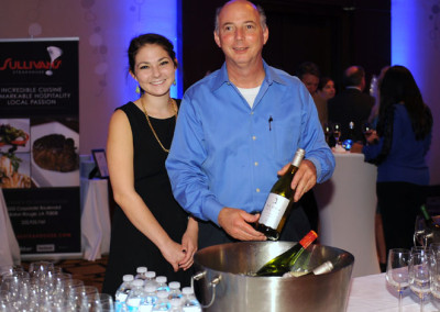 A man and a woman stand behind a promotional booth at an event, smiling, with a bottle of wine and a bucket of water bottles.