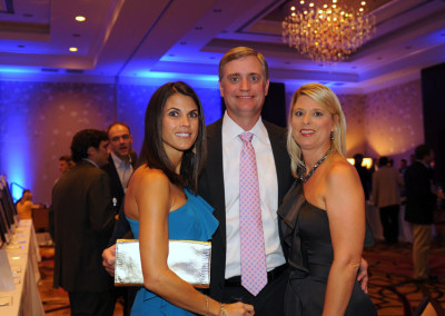 Three adults at a formal event, posing for a photo with a chandelier and blue-lit background. two women flank a man, all dressed in evening attire.