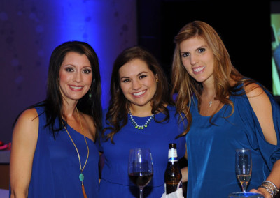 Three women in blue dresses smiling at a gala event, standing around a table with wine glasses.