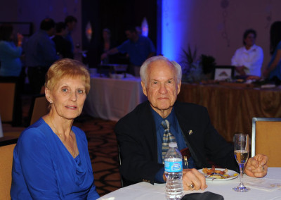 An elderly couple sitting at a table during an event, with the woman in a blue blouse and the man wearing a suit.