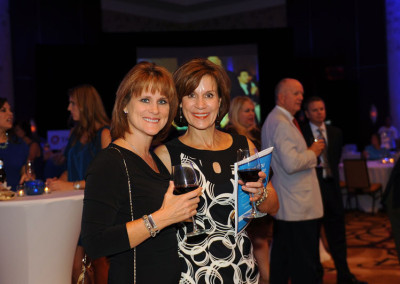 Two women smiling at a formal event, holding wine glasses with attendees in the background.